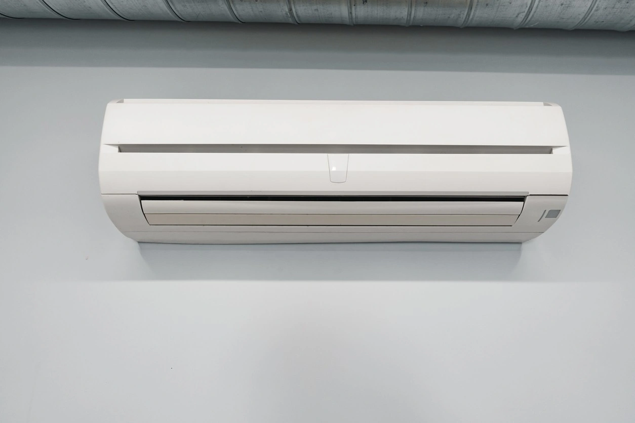 Wall-mounted AC unit on beige wall with exposed ductwork above and a TV partially visible below.