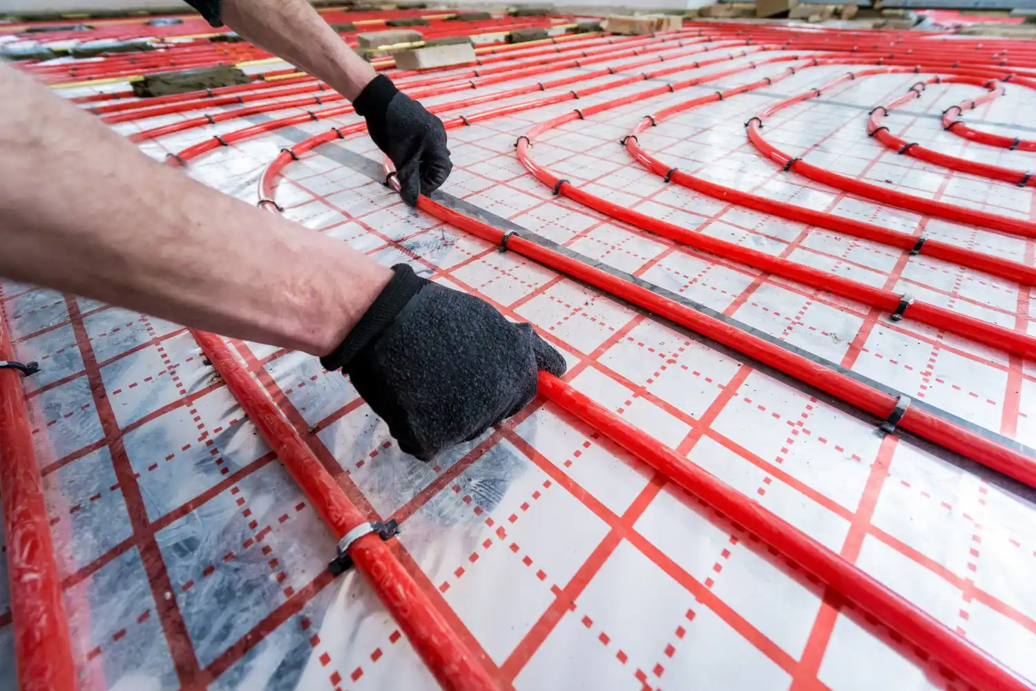 Person in black gloves secures red tubing to grid-patterned insulation layer during radiant floor heating installation.