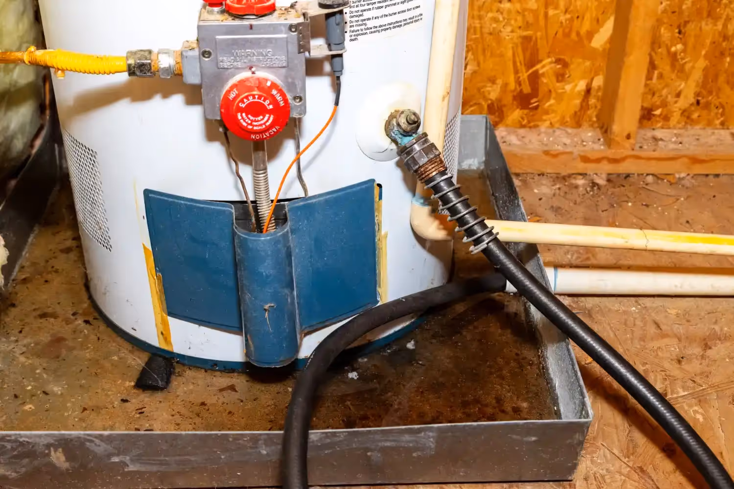 A black garden hose is connected to the drain valve of a white gas water heater, which sits in a metal drip pan filled with water. The unit is located in a crawl space with wooden framing.