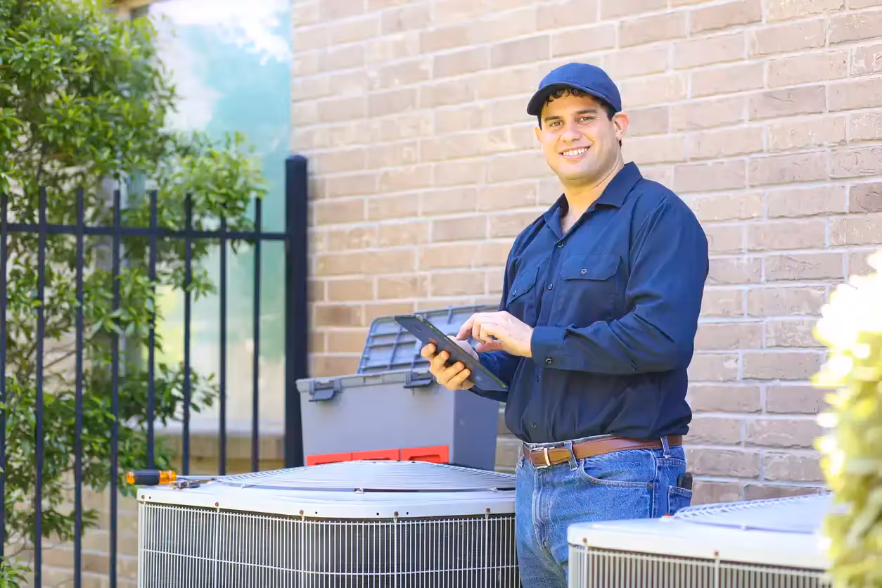 An HVAC technician in a dark blue polo and navy trousers kneels in a flowerbed to service a white air-source heat pump. He uses a handheld tool to inspect the unit's lower side panel.