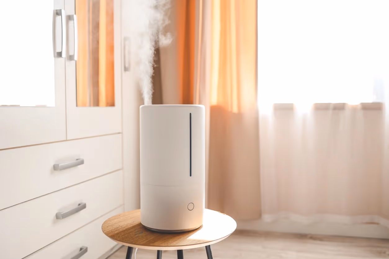 A sleek, white cylindrical humidifier sits on a small wooden table in a bright room, emitting a steady stream of cool mist. Soft sunlight filters through orange curtains in the background.