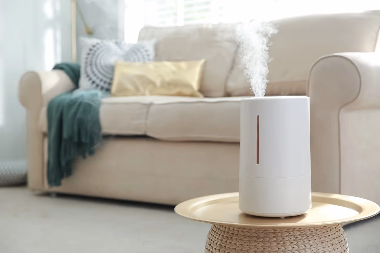 A white cylindrical humidifier sits on a gold tray atop a woven side table, releasing a plume of mist into a living room. In the background, a beige sofa is styled with decorative pillows and a green throw.