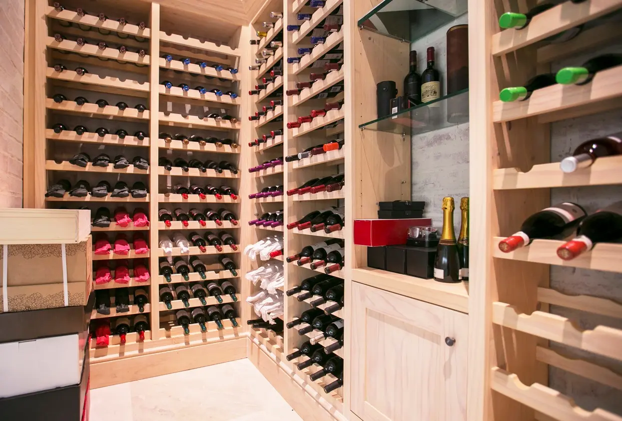 A well-lit, walk-in wine cellar features custom light-wood shelving filled with numerous wine bottles. The collection includes various labels, some wrapped in protective paper, alongside several champagne bottles