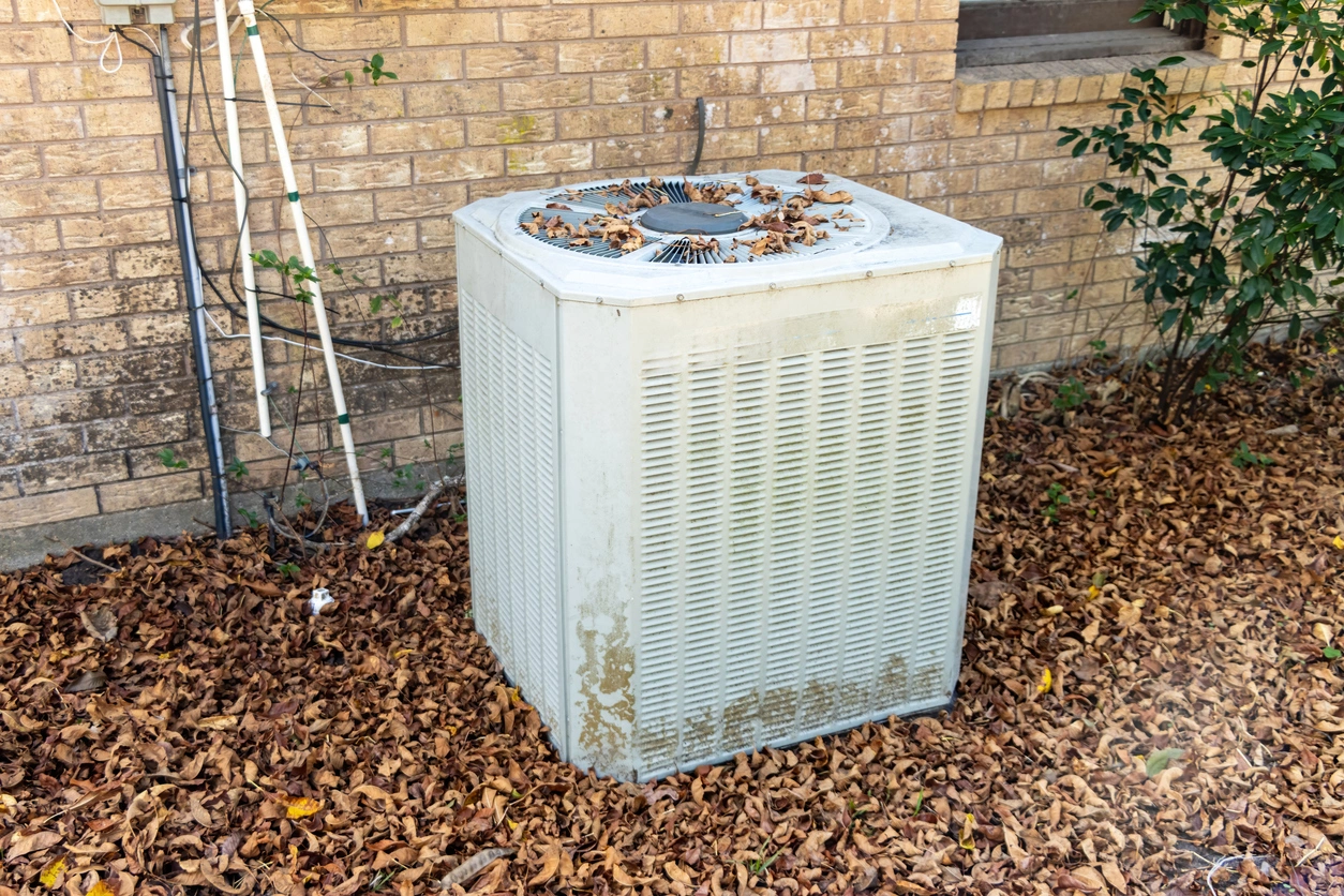 Outdoor air conditioning unit covered with fallen leaves next to a brick wall. 