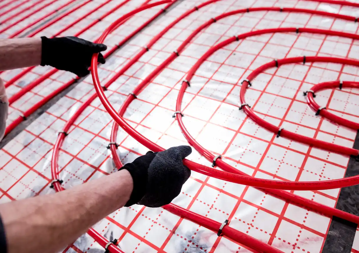 Gloved hands arrange red heating pipes onto a grid-like surface for underfloor heating installation