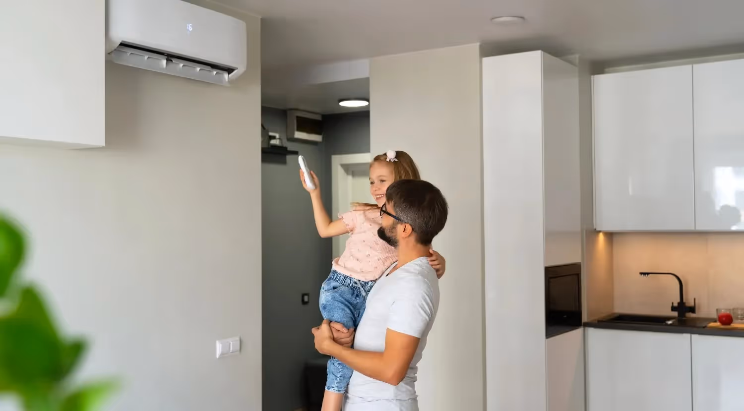 A man is joyfully holding a young girl who is pointing a remote at an air conditioner in a modern kitchen.