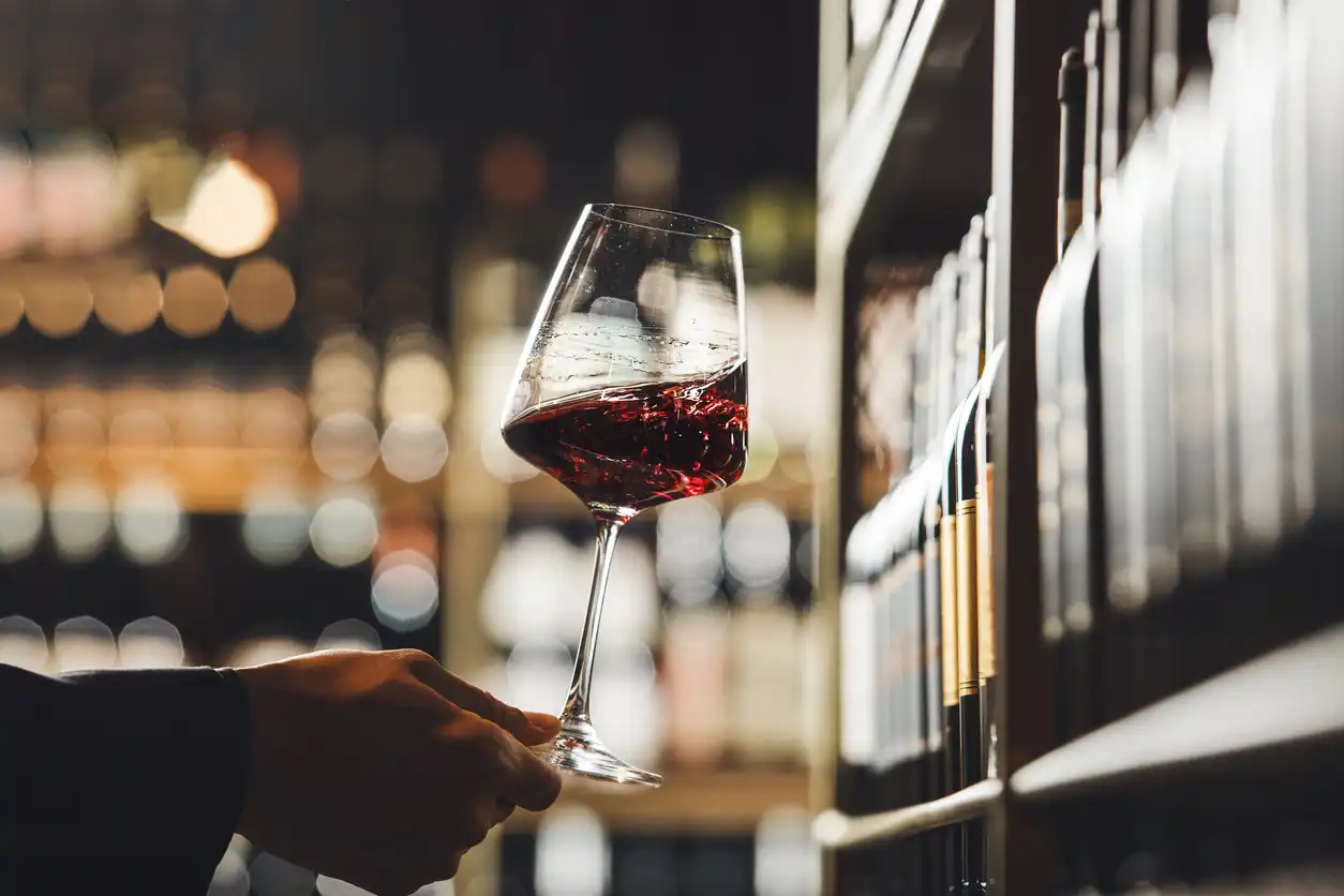 A hand holding a glass of red wine in a wine store, with shelves of wine bottles in the background. 