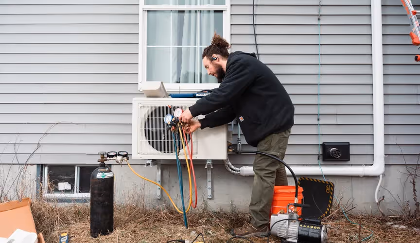 A bearded technician installs an outdoor mini-split heat pump on a gray house, connecting gauges and hoses