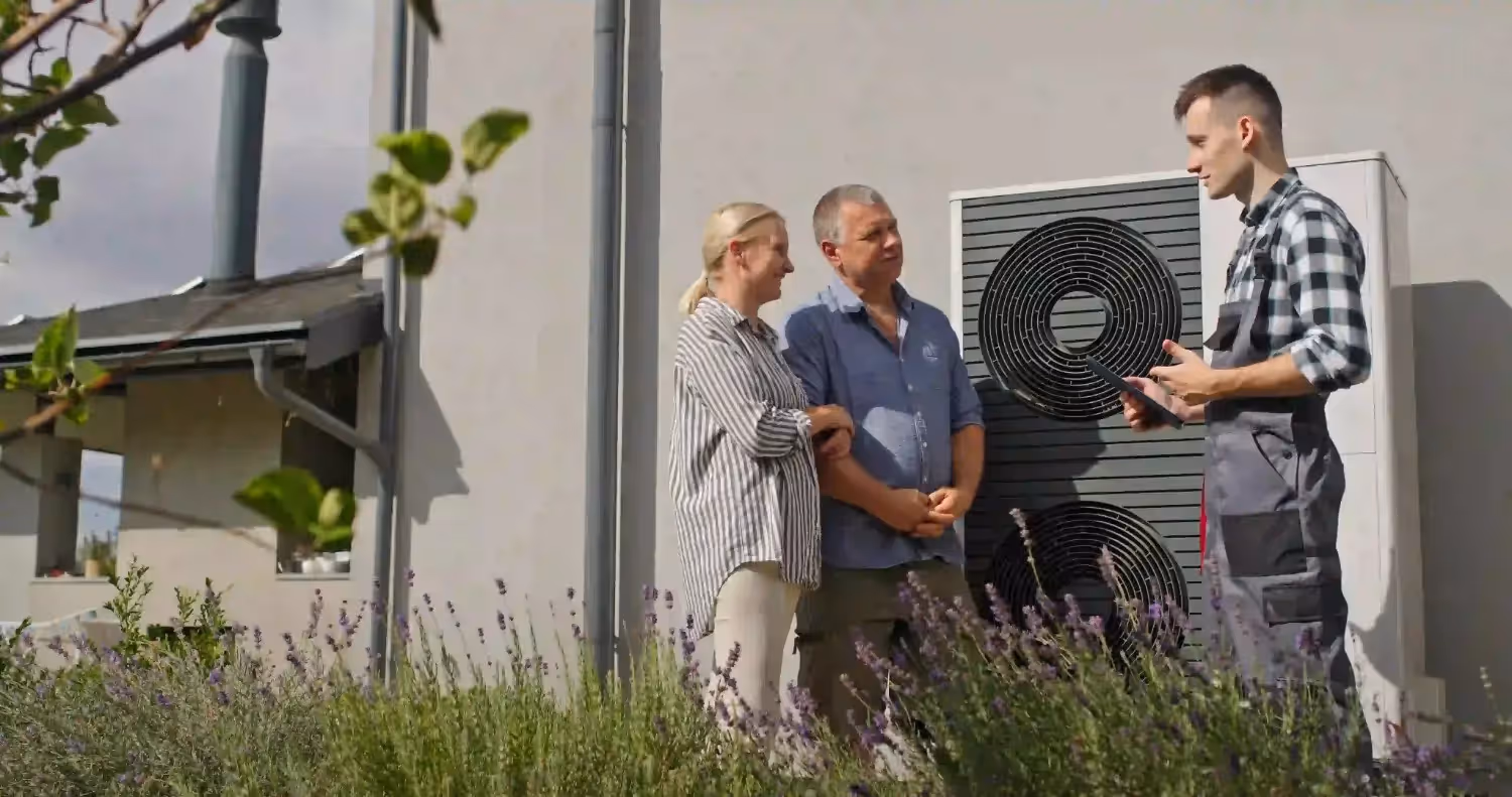 Three people stand beside large dual-fan heat pump unit outside modern home, discussing installation or service.