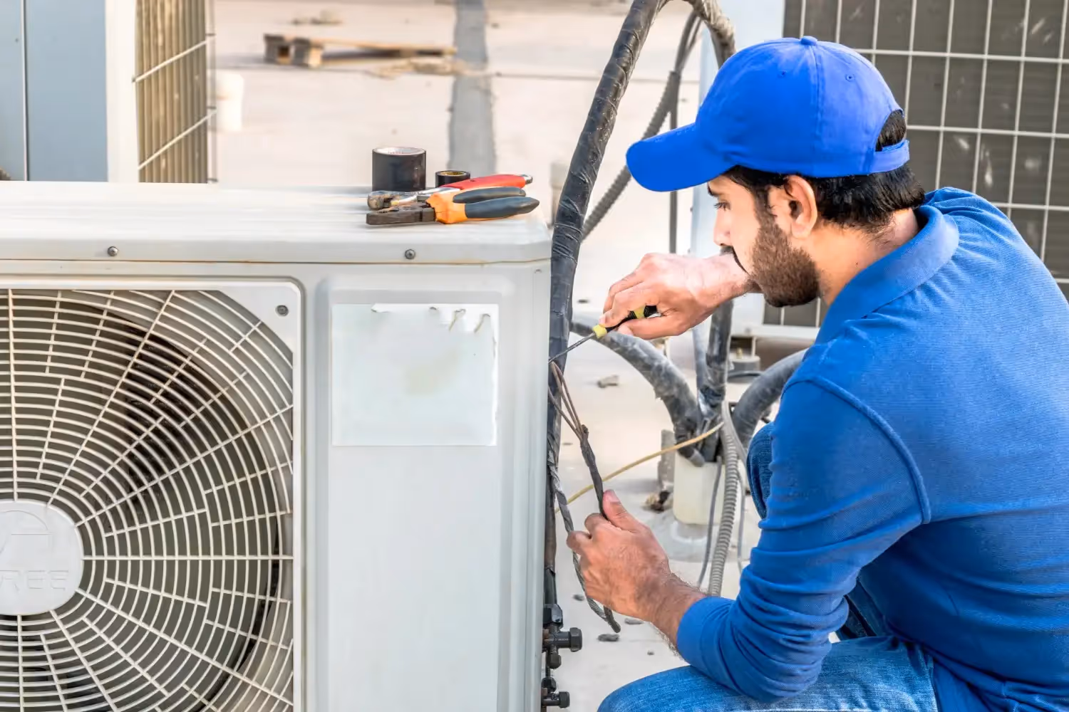 A technician in a blue shirt and cap adjusts wiring under the cover of an outdoor HVAC unit
