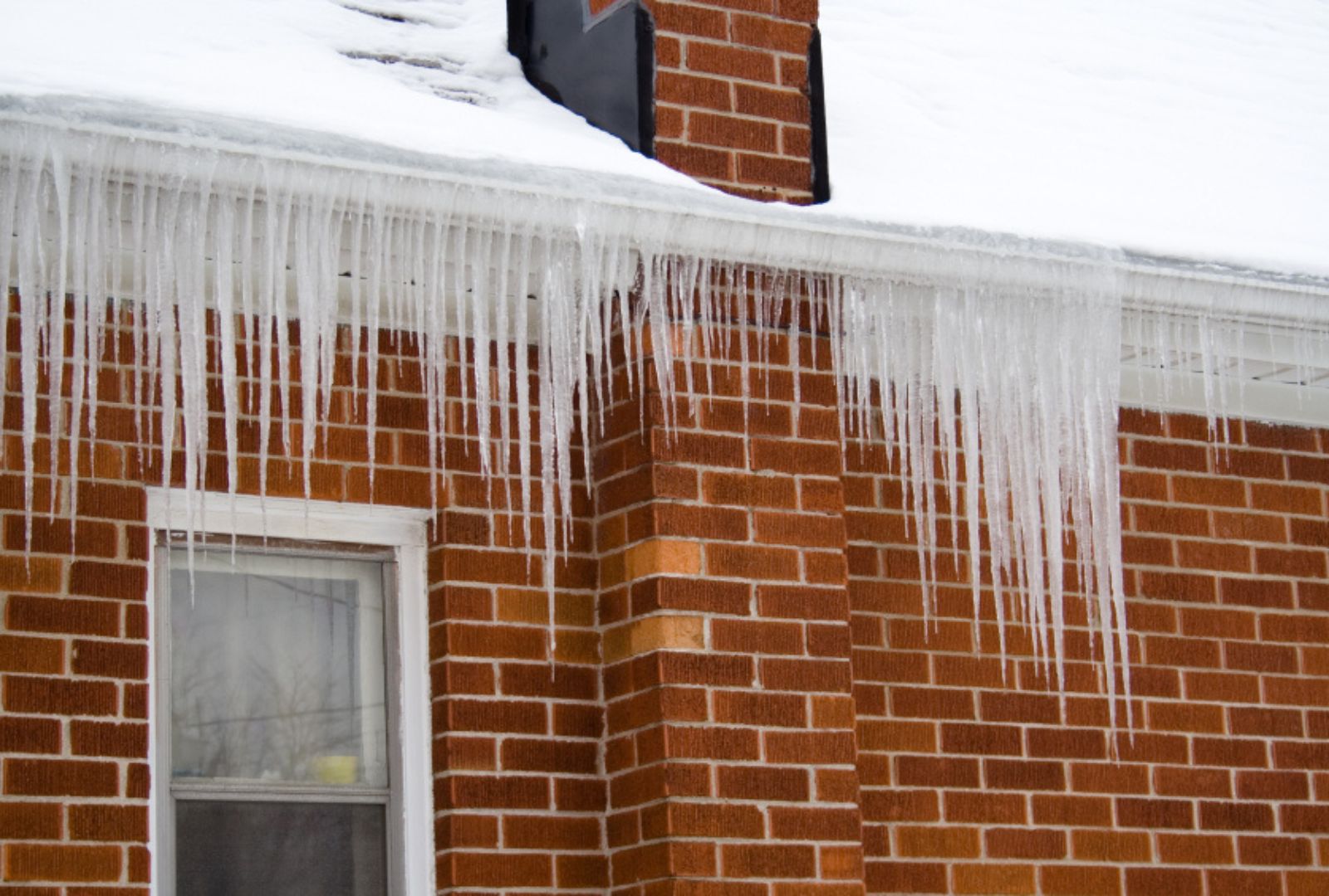 Large icicles hanging from snow covered roof of brick house indicating poor roof insulation.