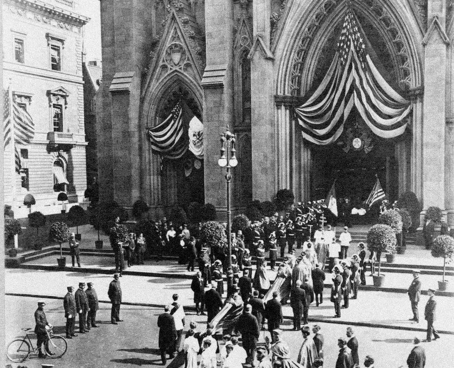 Historical black and white photo of a large crowd gathered outside a Gothic-style church with American flags draped above the entrance and people in ceremonial clothing processing towards the door.