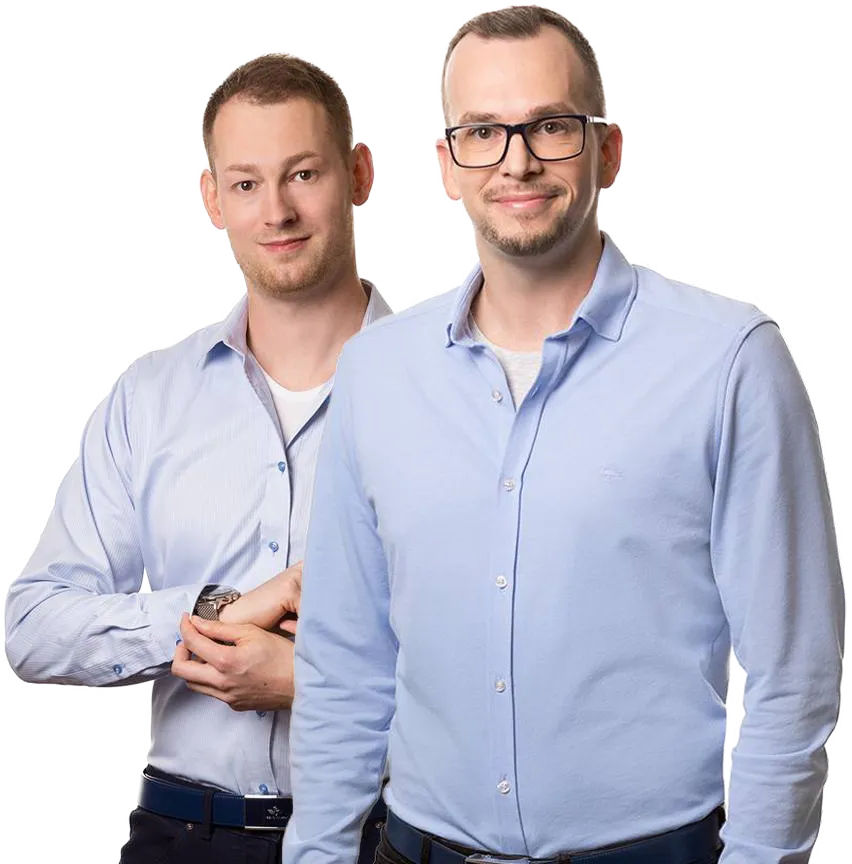 Dva muži v modrých košilích, jeden s brýlemi, stojící vedle sebe a usmívající se.