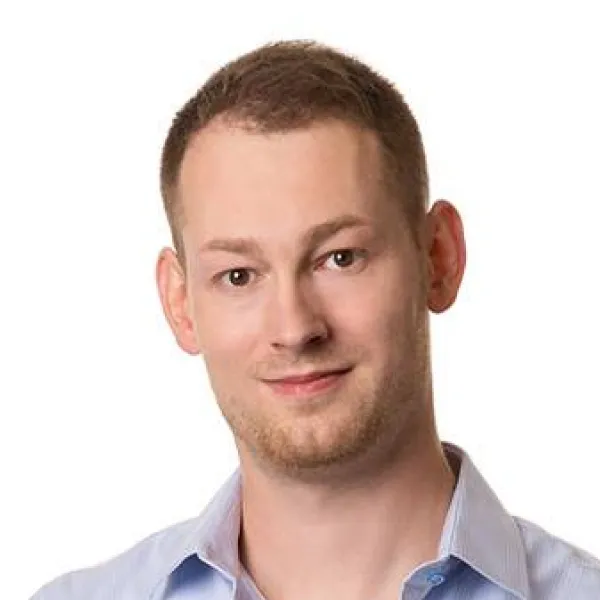 Muž s krátkými světle hnědými vlasy a jemným porostem, oblečený v modré košili, usmívá se na bílém pozadí.