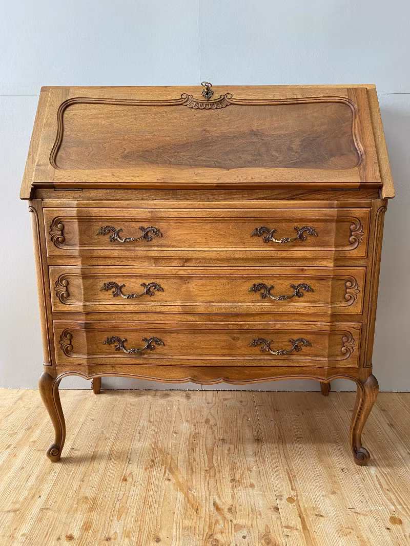 Antiker Holzsekretär mit drei verzierten Schubladen und geschwungenen Beinen auf einem Holzfußboden.