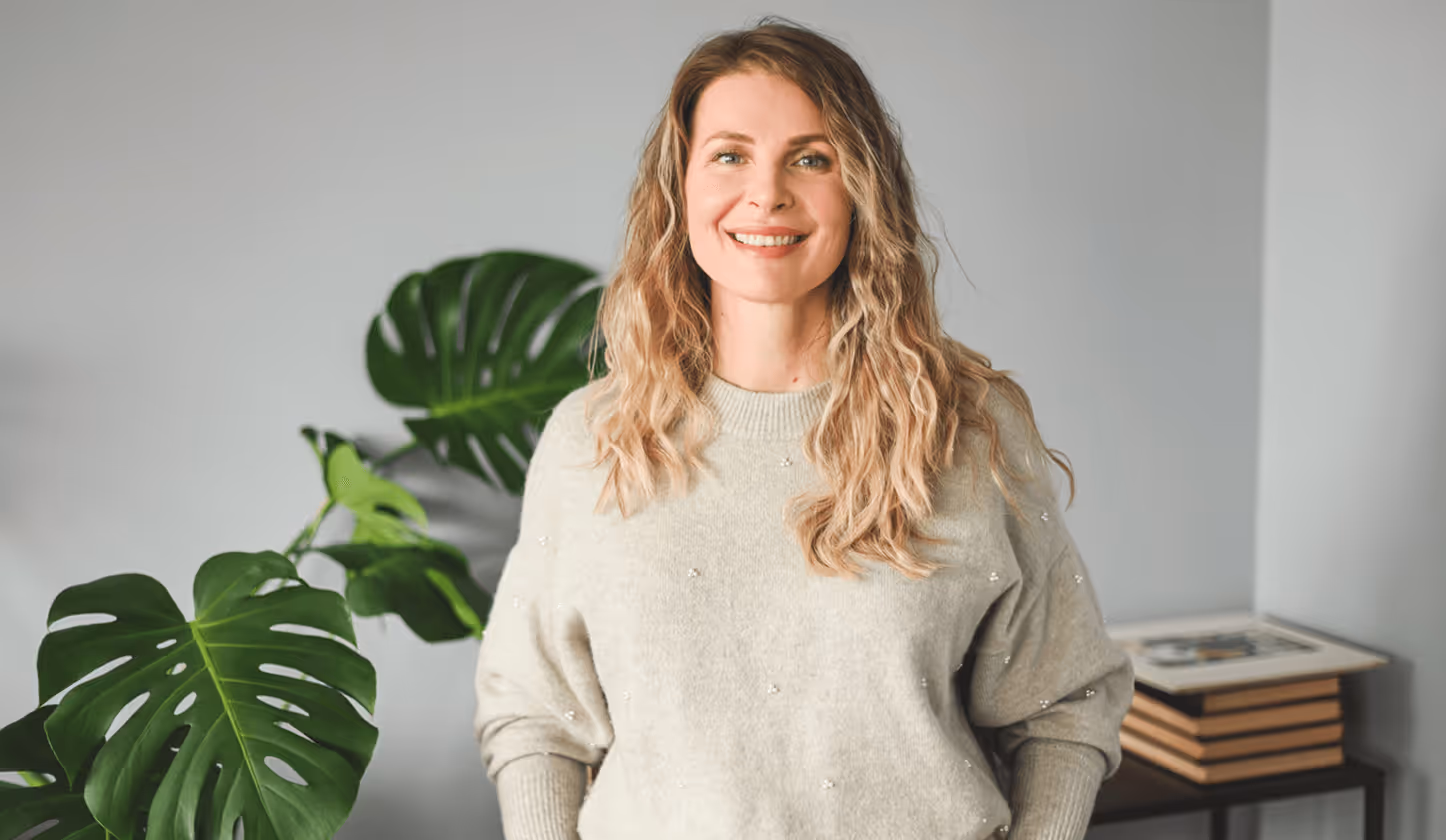 Frau mit welligem Haar in grauem Pullover lächelt, steht vor Monstera-Pflanze und Bücherregal.
