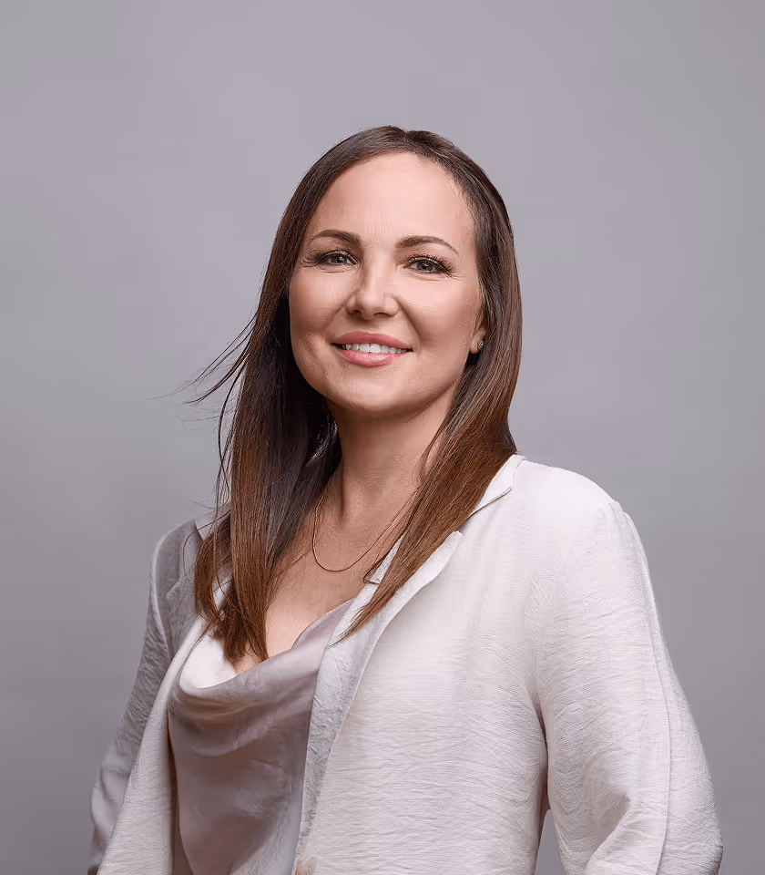 Frau mit langen braunen Haaren, lächelt in weißer Bluse vor grauem Hintergrund.