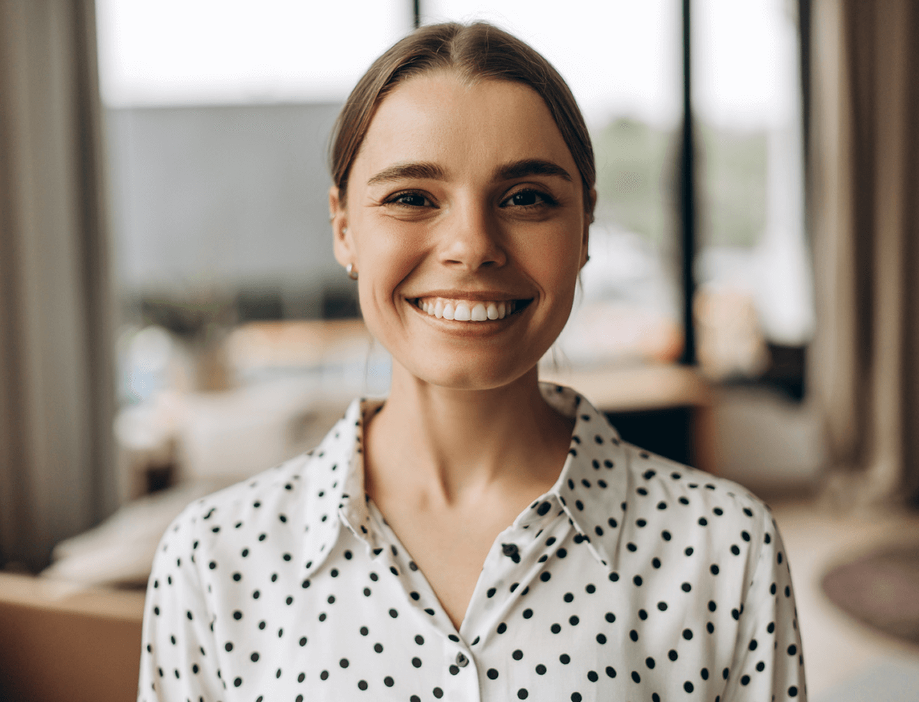 Lächelnde Frau in gepunkteter Bluse vor unscharfem Hintergrund mit Fenstern.