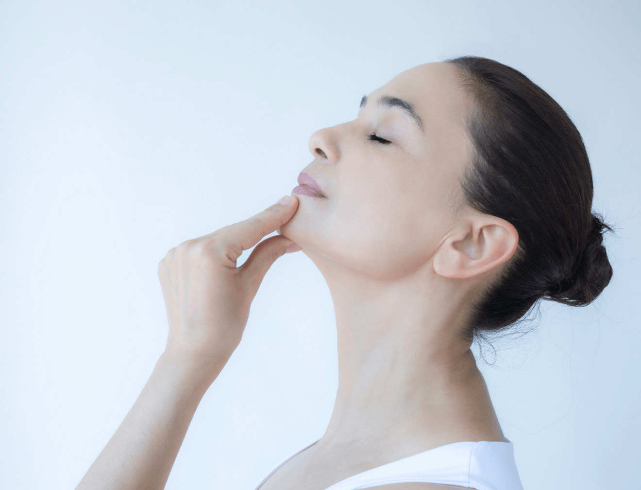 Frau in Seitenansicht hält Kinn mit geschlossenen Augen, entspannt vor weißem Hintergrund, trägt weißes Oberteil.