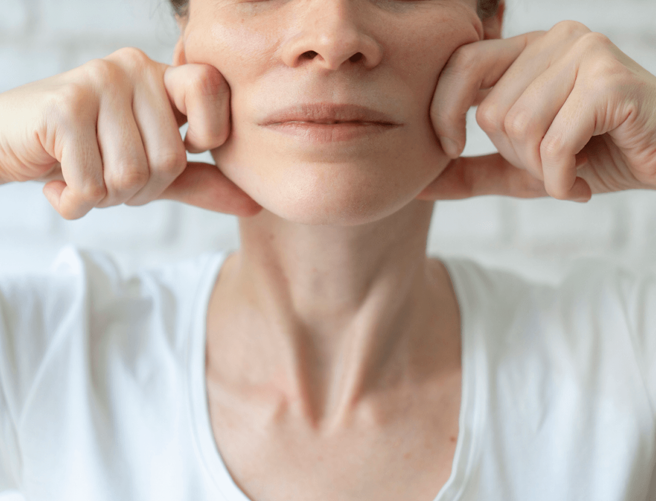 Frau in weißem T-Shirt macht Gesichtsyoga-Übung, hebt Wangen mit Fäusten vor hellem Hintergrund.