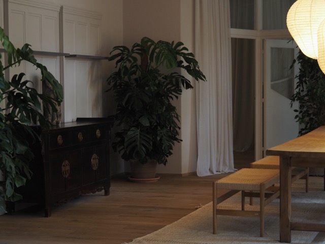 Cozy dining room with wooden table, woven benches, large leafy plants, and hanging paper lanterns.