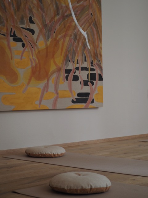 Two meditation cushions on mats on a wooden floor under an abstract orange and brown painting.
