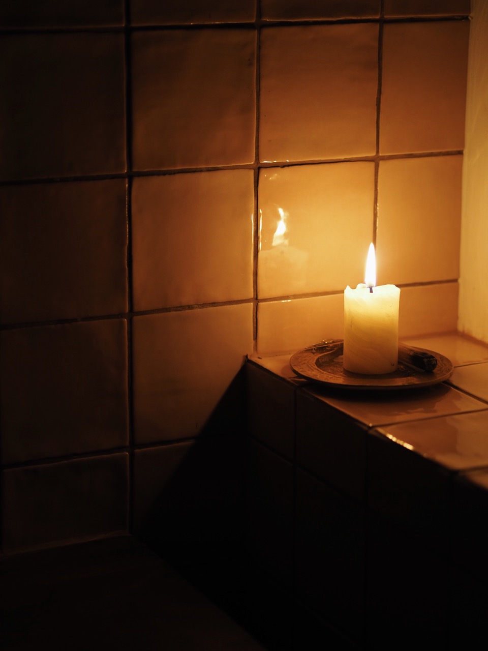 Lit white candle on a brass plate in a corner with orange tiled walls dimly lit by the flame.