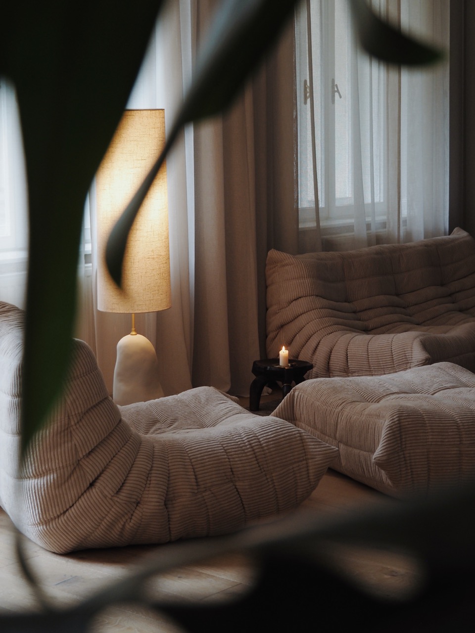 Cozy living room with beige tufted sofas, warm floor lamp, sheer curtains, and a lit candle on a small table.