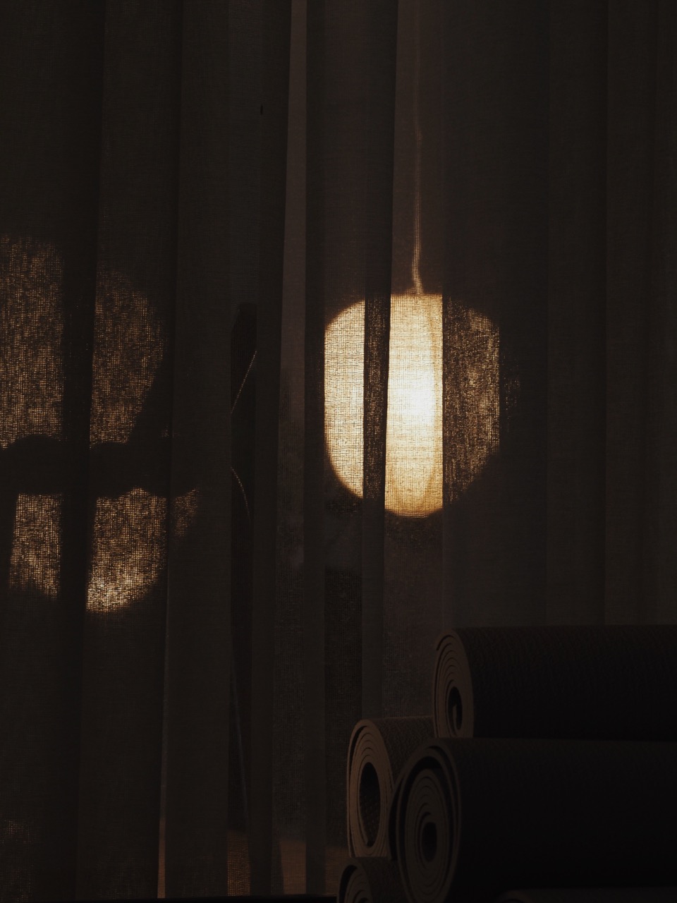 Dimly lit room with sheer curtains, a glowing round lamp behind, and rolled yoga mats in front.