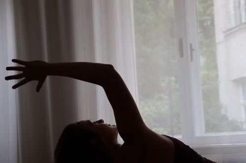 Silhouette of a person stretching an arm upward against a window with sheer curtains.