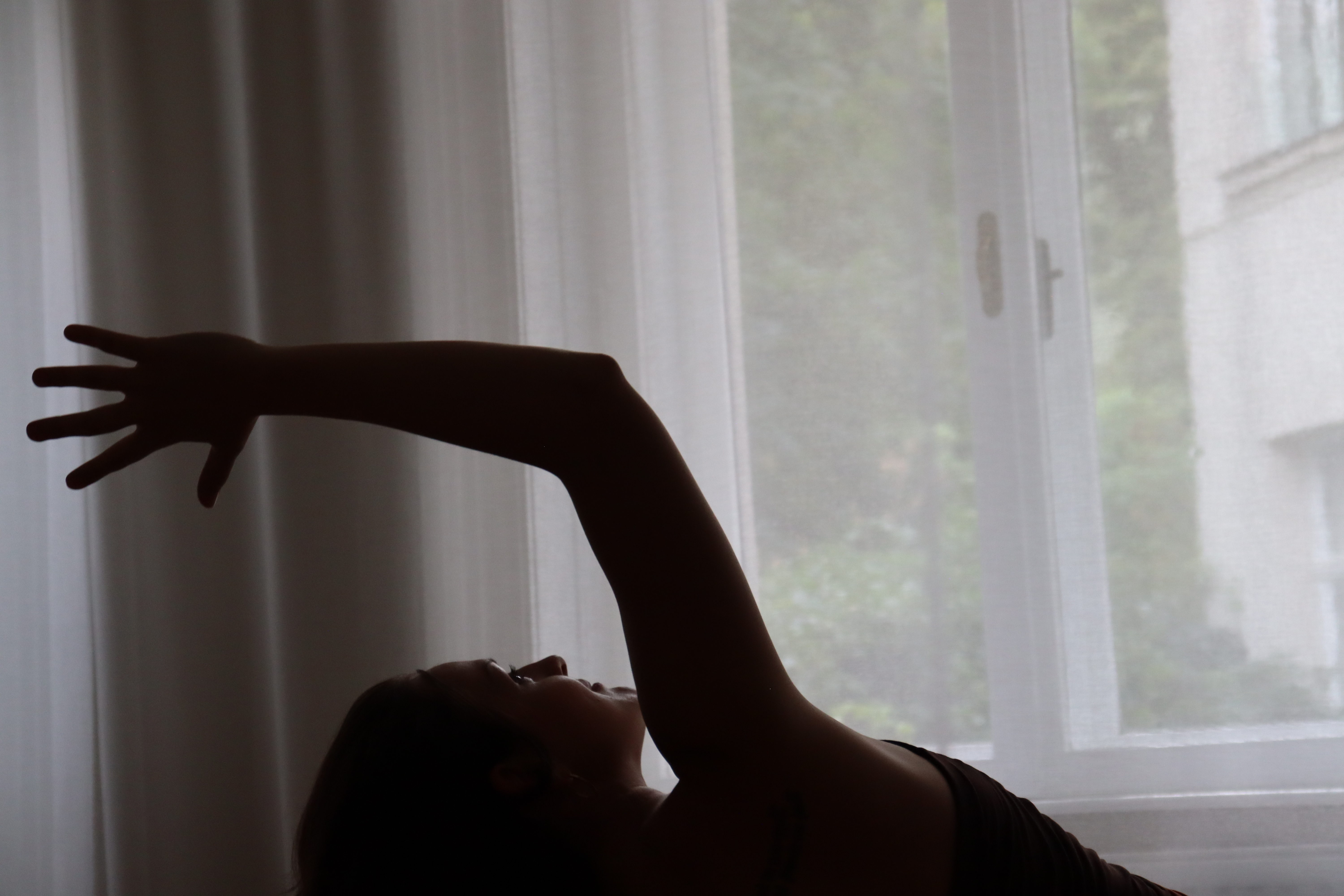 Silhouette of a person stretching an arm upward against a window with sheer curtains.