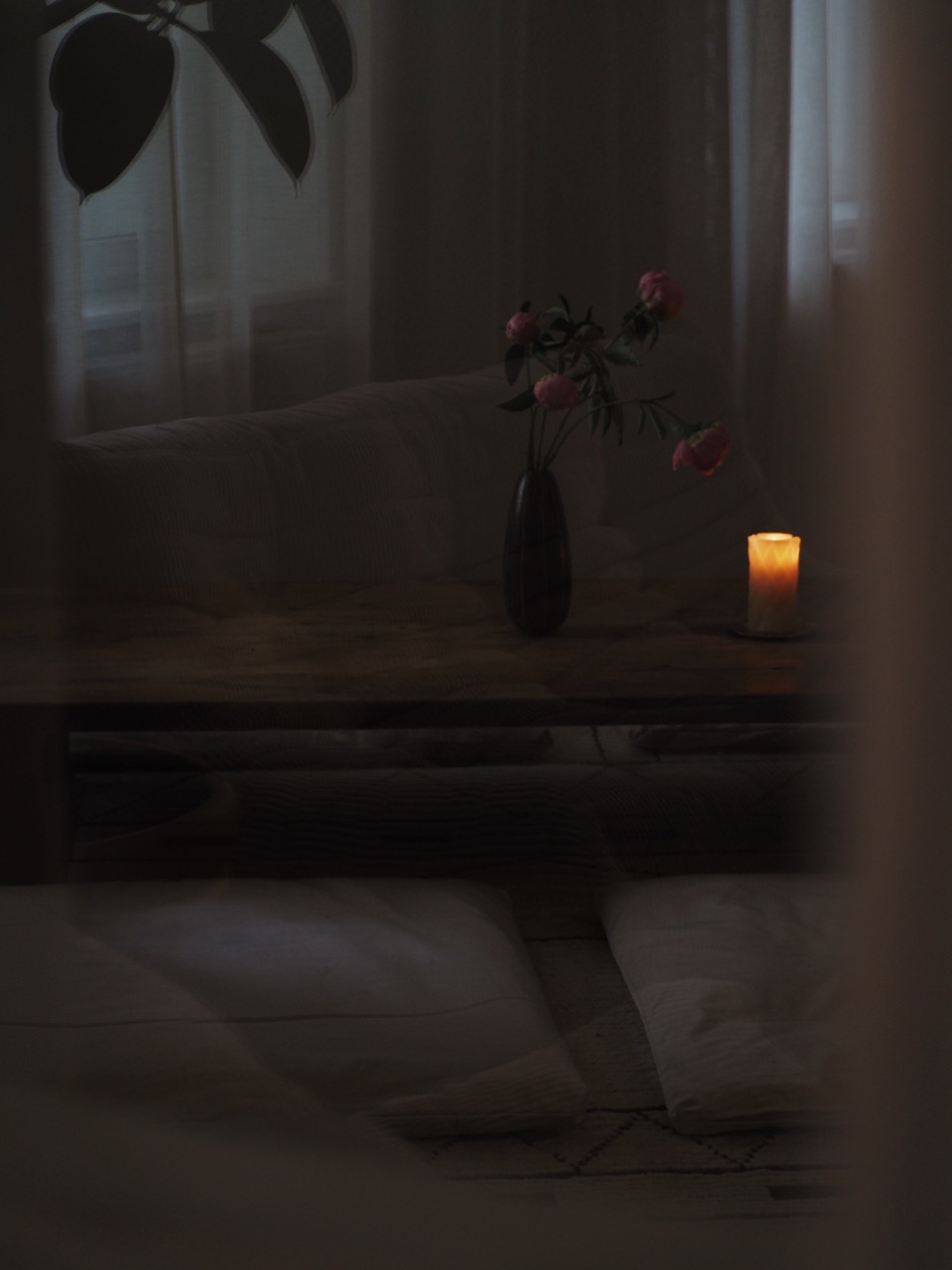 Dimly lit cozy room with wooden table, vase of pink flowers, lit candle, cushions, and sheer curtains.