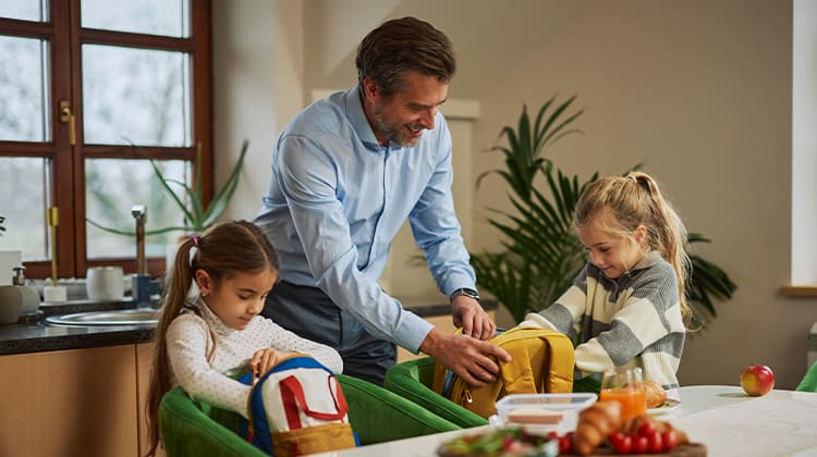 Father and his two young daughters unpack backpacks together at the kitchen table.