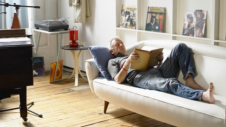 A man reads a book while lying on the couch in a room where several household items have been left out rather than put away.