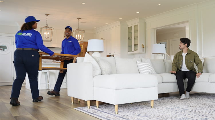 Two 1-800-GOT-JUNK? Truck Team Members lifting a table out of a living room while a customer relaxes on the couch.