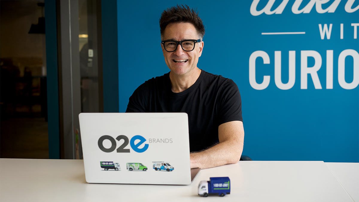 Brian Scudamore sitting at an open office desk with a laptop and a small 1-800-GOT-JUNK? toy truck in front of him