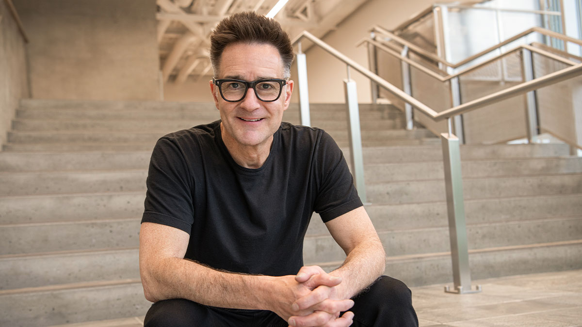 Brian Scudamore wearing glasses, smiling and sitting down in a grey cement stairwell.