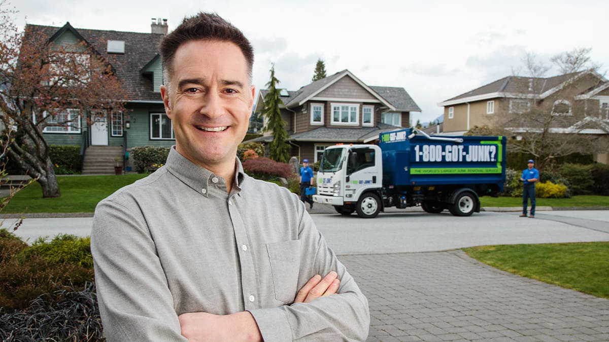 Brian Scudamore smiling and standing on a driveway in a neighborhood with a 1-800-GOT-JUNK? truck in the background with two truck team members standing around the truck.