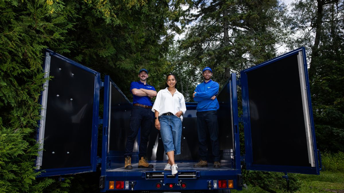 Two blue-uniformed 1-800-GOT-JUNK? team members standing in the back of an open-door junk removal truck alongside a client in a white shirt and denim jeans.