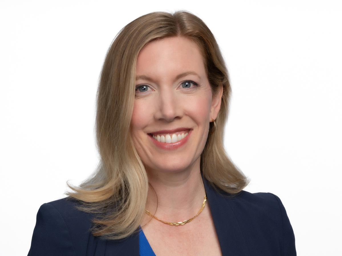 Smiling woman with blonde hair wearing a blue top, navy blazer, gold necklace, and earrings.