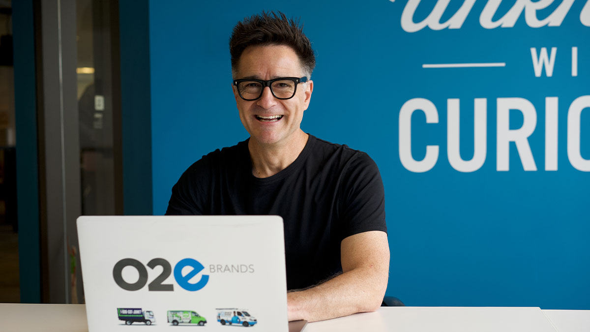 1-800-GOT-JUNK? founder Brian Scudamore smiling at a desk behind an O2E Brands laptop.