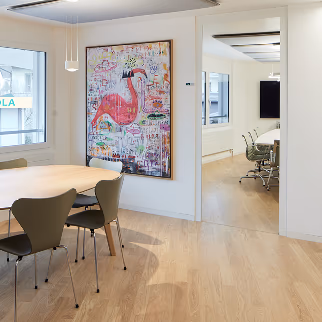 Modernes Besprechungszimmer mit rundem Holztisch, grünen Stühlen und einem großen farbenfrohen Flamingo-Gemälde an der Wand.