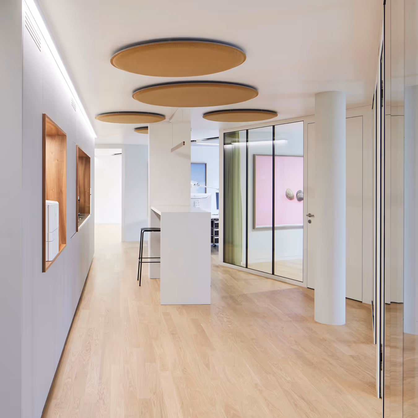 Moderner heller Flur mit Holzboden, weißen Wänden, runden Deckenpaneelen und Glasabtrennung zu einem Raum mit Kunst an der Wand.