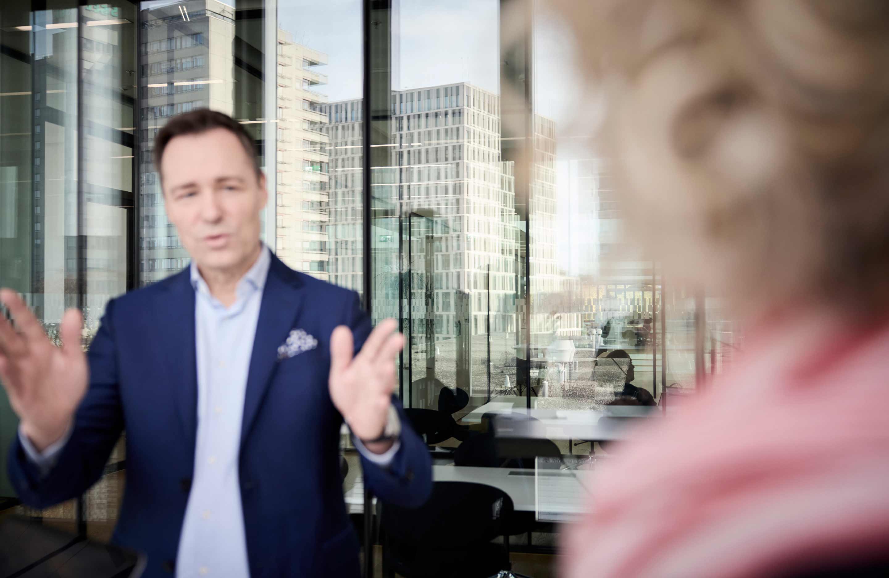 Mann im dunkelblauen Anzug spricht mit verschwommener Person im Vordergrund in einem modernen Bürogebäude mit großen Fenstern und Blick auf Hochhäuser.