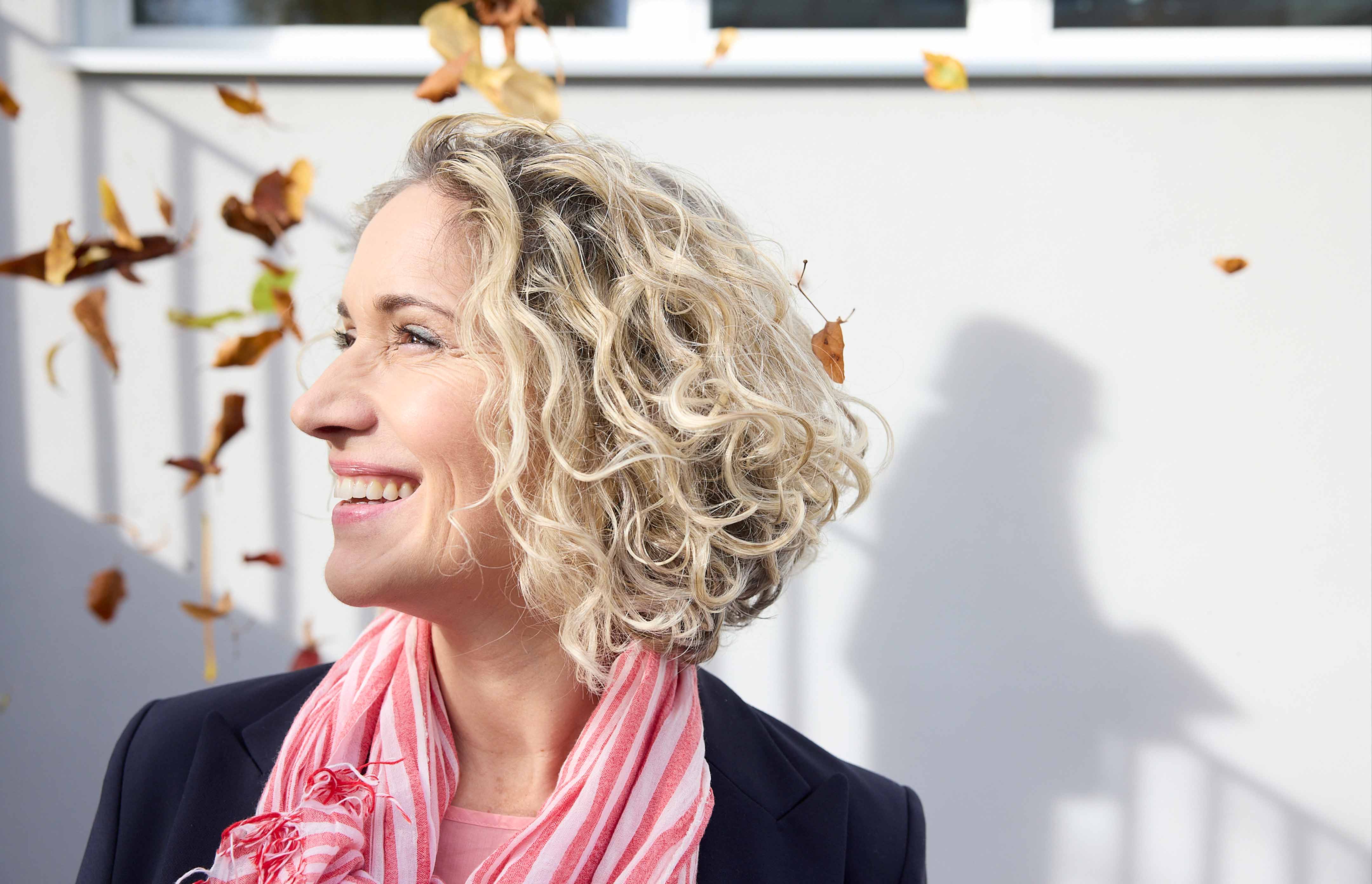 Frau mit lockigem blondem Haar, die lächelnd zur Seite schaut, während Herbstblätter um sie herum fliegen.