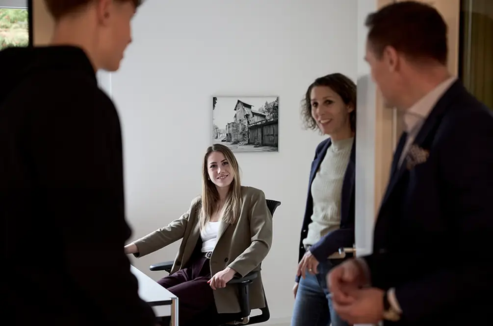 Drei Personen in einem Büro, eine Frau sitzt am Schreibtisch und lächelt, zwei andere stehen und unterhalten sich.