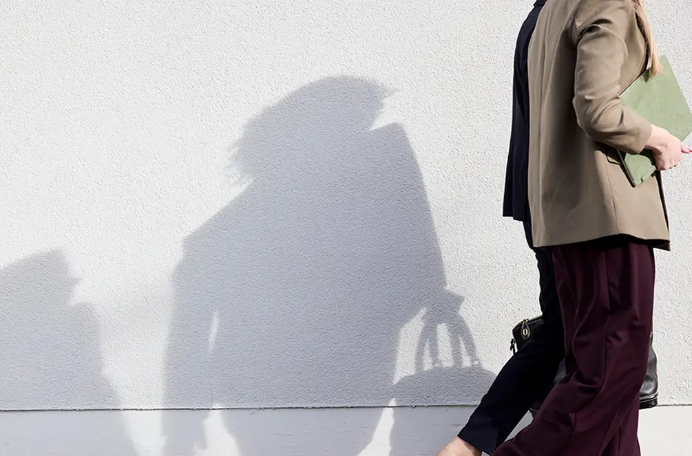 Schatten von zwei gehenden Personen an einer hellen Wand, eine hält eine Tasche, die andere eine Mappe.