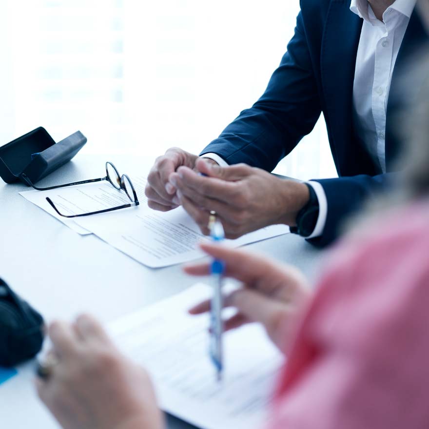 Zwei Personen am hellen Sitzungstisch mit Dokumenten, eine Person trägt einen Anzug und sitzt mit verschränkten Händen, die andere hält einen Stift.