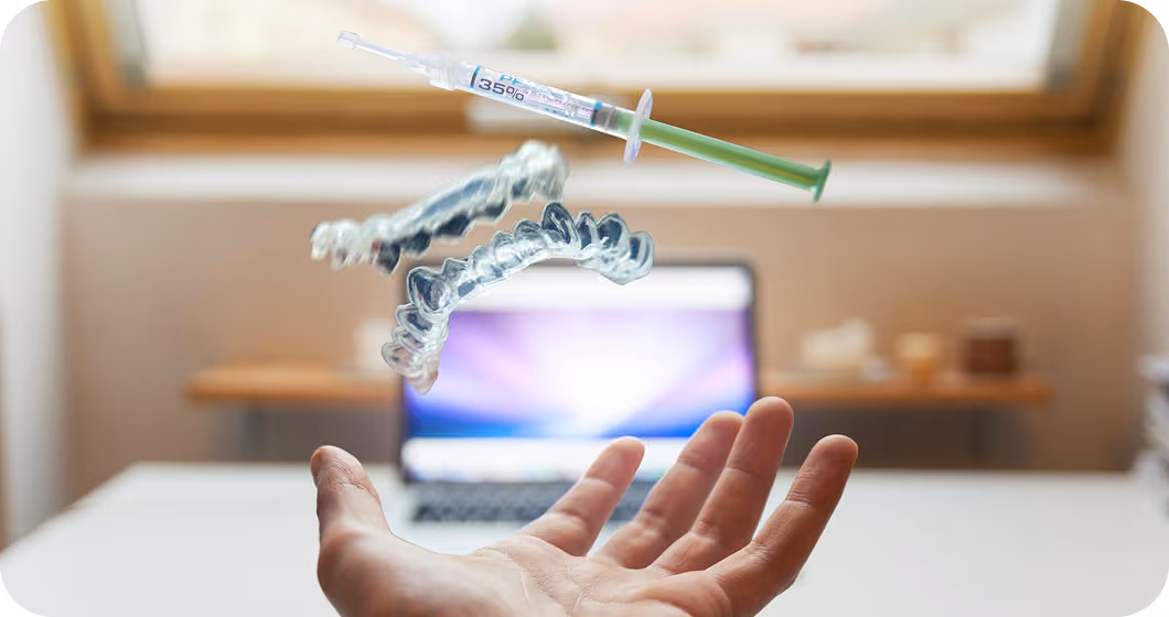 Hand with a floating dental aligner and a syringe with green liquid in front of a blurred laptop screen.