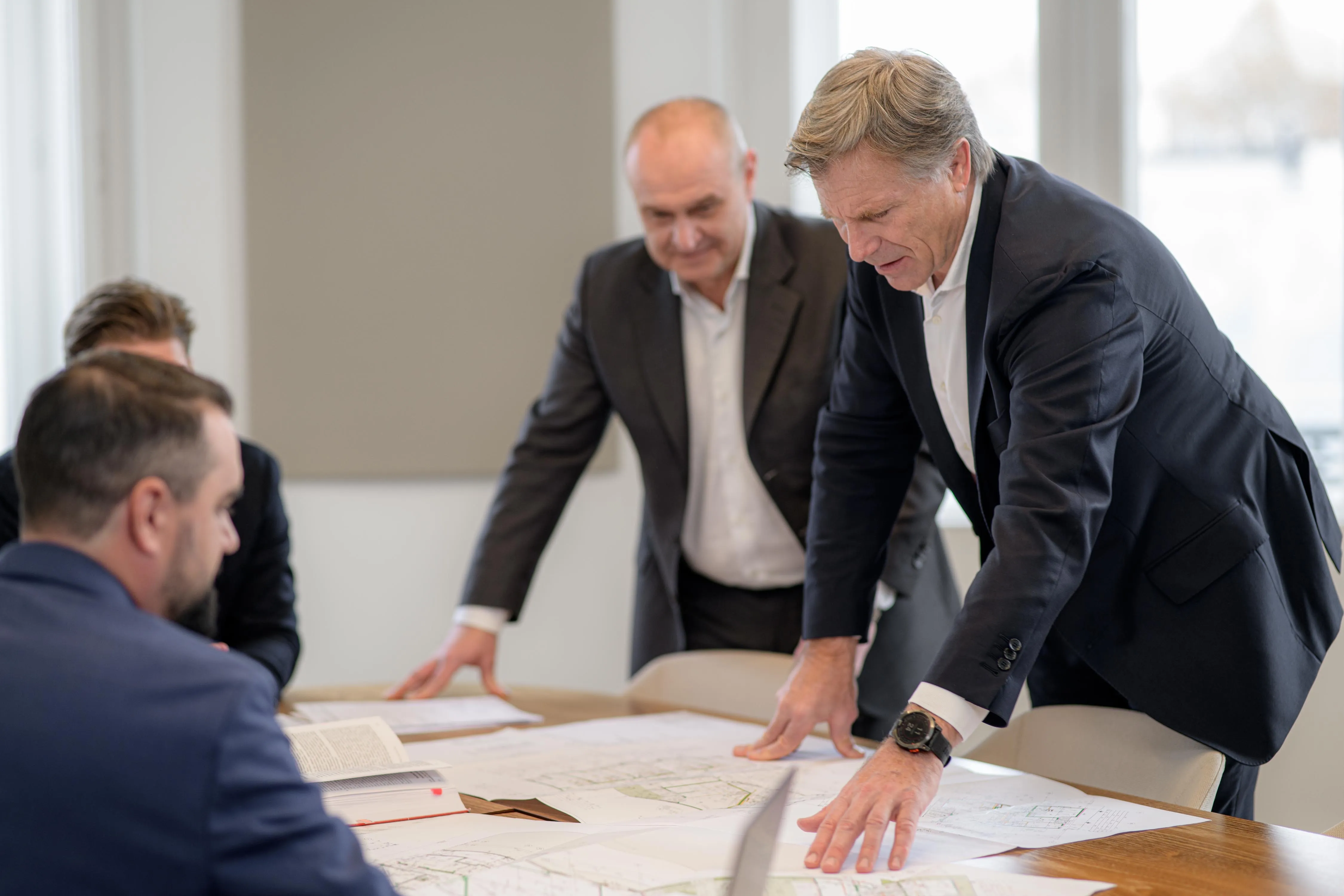 Vier Geschäftsleute besprechen Pläne und Dokumente auf einem Tisch in einem modernen Büro mit großen Fenstern.